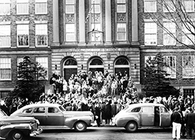 A large group of students, boys and girls, and faculty in light coats and jackets, stand outside the main entrance of St. George High School.  Three cars are pulling up to the curb and dropping off more students and faculty.